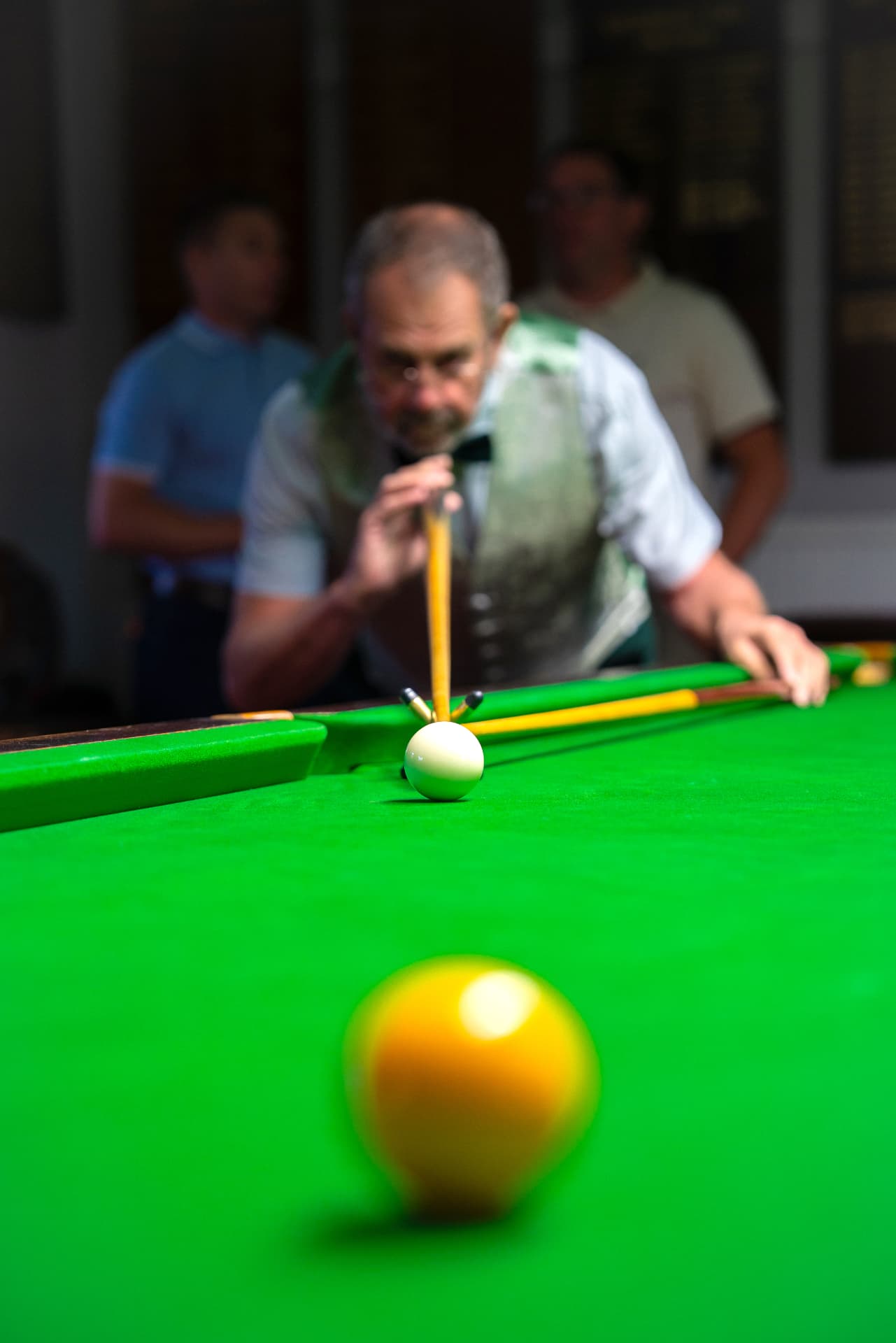 A member down on the cue playing a shot, with others watching from behind the table.