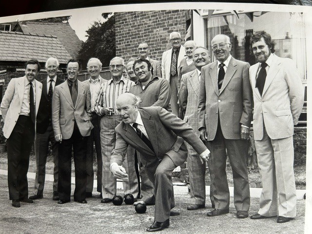 A black-and-white archive photograph, 1970s: a group of club members in front of the clubhouse, one gentleman in the foreground mid-bowl, Les Dawson standing in the back row.